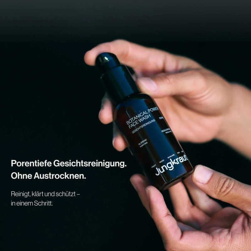 Zwei Hände halten eine braune Flasche des Jungkraut Botanical Power Face Wash. Links im Bild steht der Text: „Porentiefe Gesichtsreinigung. Ohne Austrocknen. Reinigt, klärt und schützt – in einem Schritt.“ Der Hintergrund ist dunkel und lenkt den Fokus auf das Produkt.